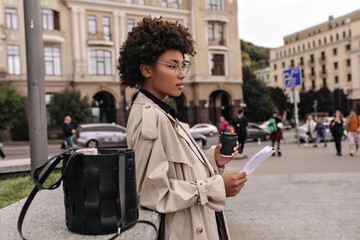 Profile portrait of curly dark-skinned woman holding paper sheet and coffee cup. Attractive lady in beige trench coat leans on bordure.