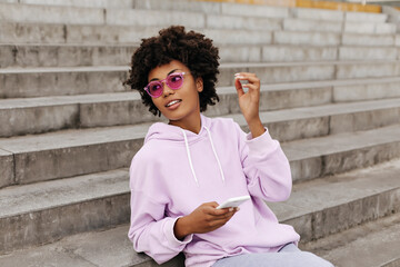 Charming young brunette curly dark-skinned woman in purple hoodie and bright pink sunglasses sits on stairs outdoors and holds white phone.