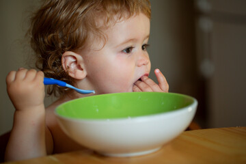 Funny baby eating food himself with a spoon on kitchen.