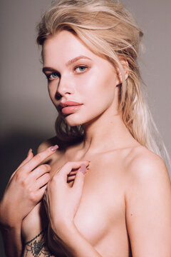 Closeup of pretty young woman face with blue eyes, blonde hair, has nude makeup, perfect soft skin, standing shirtless with bare shoulders, looking at camera. Studio grey background