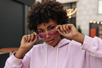 Stylish young dark-skinned woman in pink hoodie and colorful stylish sunglasses looks into camera and poses outdoors.