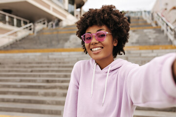 Happy cheerful curly dark-skinned woman in purple oversized hoodie and pink sunglasses smiles and takes selfie outside near stairs.