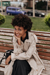 Happy excited brunette woman sits on wooden bench. Cheerful girl in beige trench coat and eyeglasses smiles sincerely.