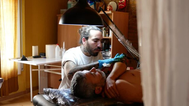 Panning scene of a tattoo artist working with a client in his studio.
