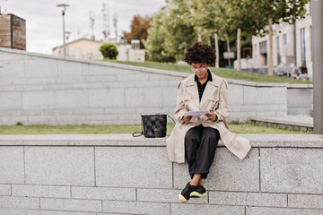 Stylish young brunette dark-skinned woman in black pants and beige trench coat sits outside and holds paper sheet.