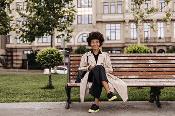 Joyful brunette woman sits on wooden bench outside. Good-humored curly dark-skinned lady in black pants and beige trench coat smiles.