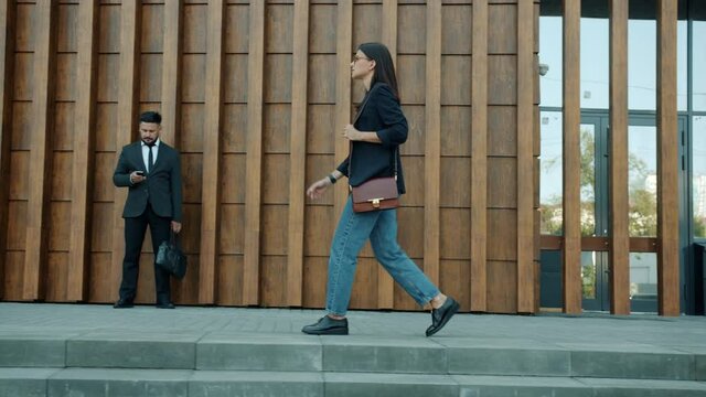 Side view of stylish young woman entrepreneur walking along modern office building going to work alone while people are moving in background.