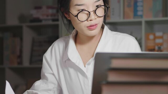Asian woman wearing glasses working at night, finding documents and looking at laptop, rechecking information. Working hard at home, working from home concept