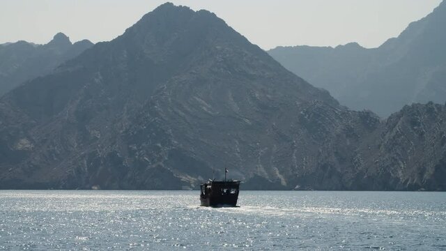 Wide shot of traditional wooden dhow boat sailing towards the tall and dramatic fjord cliffs in the glassy blue ocean water. The sunlight sparkles on the water in Musandam, Oman.