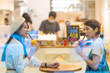 Fototapeta premium Young Asian woman friends sitting by the window in cafe with drinking fruit juice and talking together with happiness. Attractive female friendship enjoy leisure activity and meeting in the city