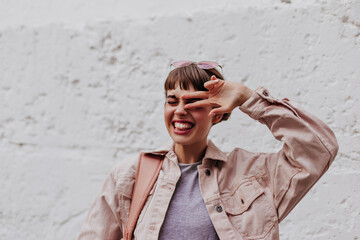 Happy woman with brunette hair showing peace sign on white backdrop. Excited short haired lady in light outfit makes funny face on background of wall..