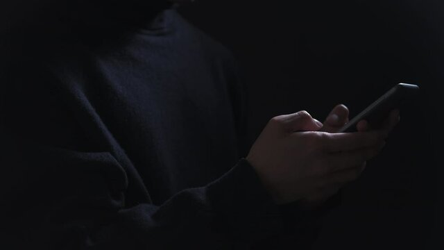 Mobile hacker attack. Online scam. Cyber crime. Internet fraud. Unknown spy man in black using phone texting password information isolated on dark.