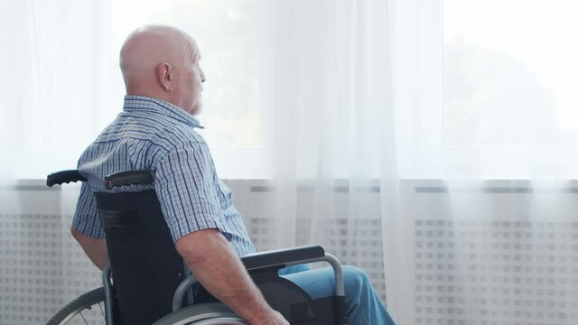 Disabled old man is sitting in a wheelchair at home alone. A handicapped person in a wheelchair is looking out the window. Paralysis and disability.