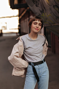 Hipster girl with short hairstyle posing at street. Brunette woman in denim jacket, grey t-shirt and jeans with black belt smiling outdoors..