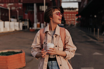 Cheerful woman with cup of coffee smiling in city. Lady with short hair in pink sunglasses, beige denim jacket and jeans posing outside..