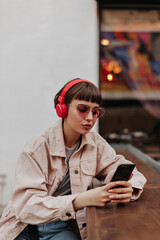 Fototapeta premium Modern girl in red headphones chatting in cafe. Short-haired lady in pink glasses and denim beige outfit listening to music outside..