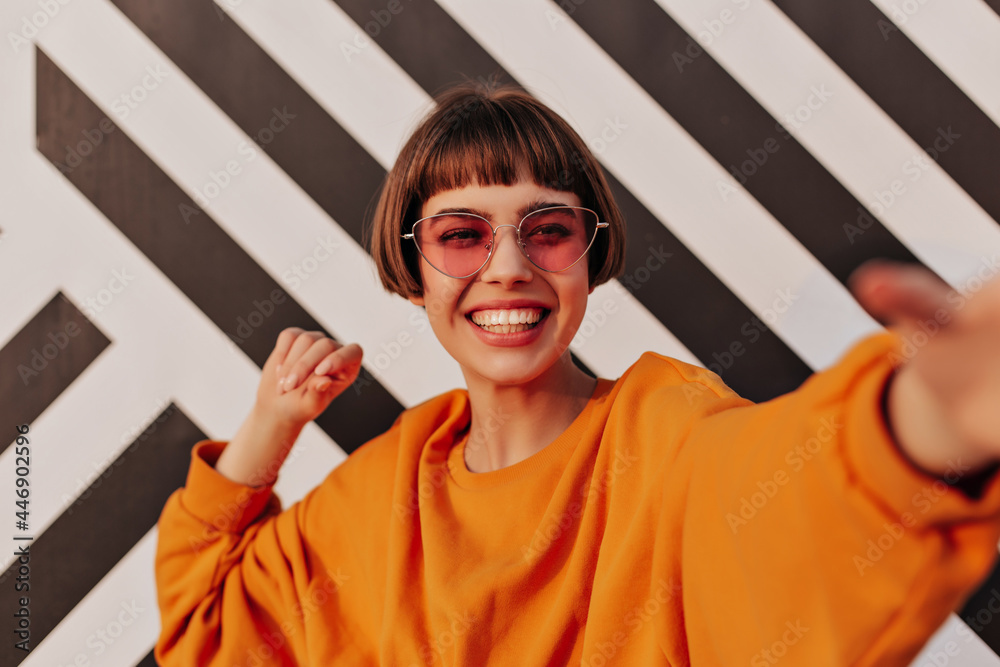 Wall mural Cheerful short-haired girl in bright clothes smiling outside. Modern woman in sunglasses making selfie on striped backdrop.. - Wall murals