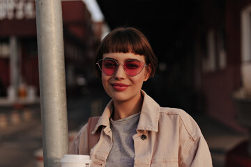 Short-haired girl in pink sunglasses smiling outdoors. Happy woman with brunette hair in beige cool jacket and grey t-shirt posing in city..