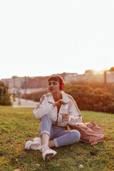 Short-haired woman in jeans and white jacket sits on grass, opens water bottle and listens to music in headphones outdoors.