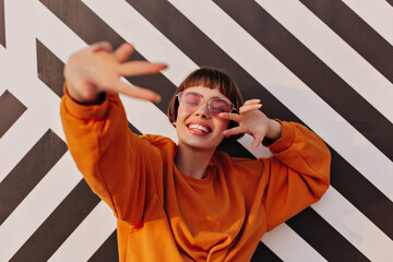 Cheerful lady in pink sunglasses smiling outside. Joyful woman in orange clothes showing peace sign...