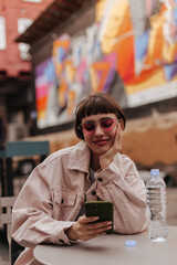 Modern lady with brunette hair chatting outdoors. Optimistic girl in denim light jacket and pink sunglasses smiling and holding phone outside..