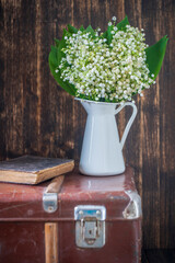 Beautiful bouquet of lilies of the valley in a white enamel vase on an old suitcase next to a book