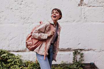 Optimistic woman with short hair laughing outside. Cheerful lady with backpack in denim jacket and...