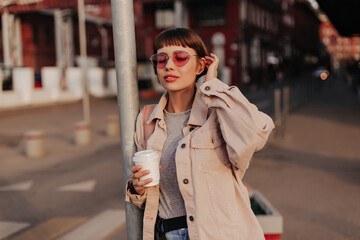 Obraz premium Fashionable lady holding cup of coffee outside. Brunette girl in denim beige jacket and pink glasses posing with closed eyes in city..