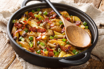 Hot rustic fried potatoes with green onions, mushrooms and bacon close-up in a frying pan on a wooden background. horizontal