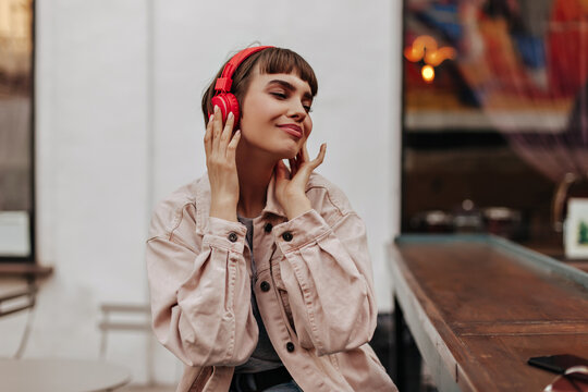 Charming brunette lady listening to music outside. Stylish girl with short hair in red headphones and beige denim outfit smiling outdoors..