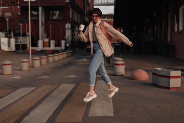 Cool woman in beige jacket jumping at street. Girl with short hair in jeans, glasses and light...