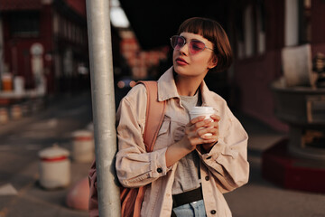 Stylish woman in tight outfit holding cup of tea in city. Short-haired girl in pink glasses and beige jacket with backpack posing outside..