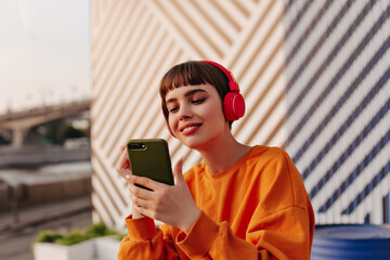 Charming lady with brunette hair in red headphones holding phone outside. Girl in orange sweatshirt...