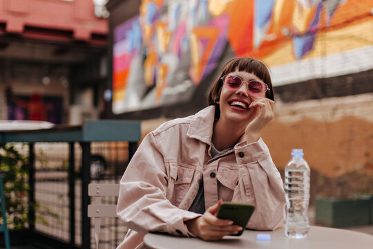 Cheerful woman with brunette hair laughing outside. Modern short-haired girl in bright glasses and denim light outfit holding phone in cafe..