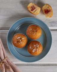 Ruddy brown fluffy muffins on a blue porcelain plate and muffin pieces with berry jam on a white wooden background