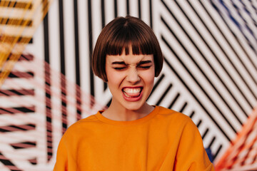 Emotional girl with brunette hair in orange sweatshirt makes funny face outside. Hipster in bright clothes showing tongue on striped backdrop..