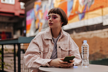 Stylish woman holding smartphone at street. Short haired teen in bright glasses and denim beige jacket posing in cafe with bottle water..