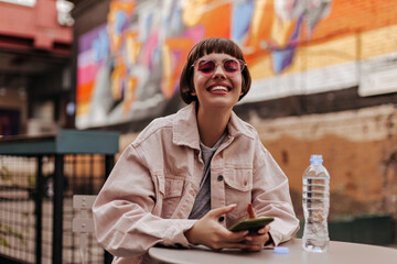 Cheerful lady with short hair smiling widely outdoor. Positive brunette girl in light jacket and pink sunglasses holding phone at street..