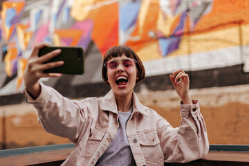 Emotional lady with short hair taking selfie outside. Brunette woman in pink glassess and light...