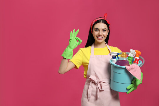 Young housewife holding bucket with cleaning supplies on pink background, space for text