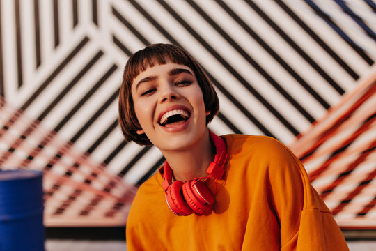 Optimistic brunette woman in orange clothes laughing in cafe. Short-haired girl with red headphones posing on striped background..