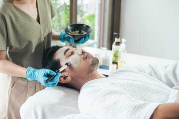 Asian men skin care. Beautician use clay mask on Asian man face. Young male relaxation in spa salon. Concept facial treatment and skin rejuvenation for men.