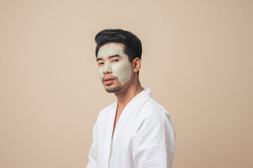 Asian man in bathrobe facial treatment and skin rejuvenation with clay mask on face in studio brown color background. - obrazy, fototapety, plakaty