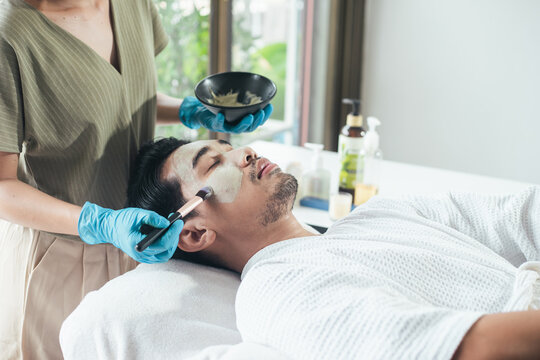 Asian men skin care. Beautician use clay mask on Asian man face. Young male relaxation in spa salon. Concept facial treatment and skin rejuvenation for men.