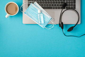Top view photo of two medical facemasks and transparent sanitizer bottle on laptop headphones and cup of drink on isolated pastel blue background with copyspace