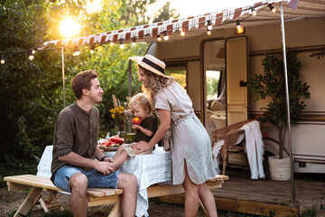 Family vacation in mobile home: young parents travel with small preschool son have picnic on terrace on sunset at rv camper trailer. People enjoy summer road voyage on caravan car. Summer outdoor trip - obrazy, fototapety, plakaty