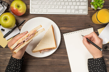 First person top view photo of hands holding sandwich over plate and writing in planner keyboard...