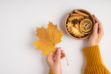 First person top view photo of woman hands in yellow sweater holding yellow autumn maple leaf and wooden bowl with cookies dried lemon slices anise and cinnamon sticks on isolated white background
