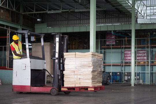 African American Man Driver Warehouse Worker In Safety Vest And Helmet On Forklift Truck In Industry Factory.