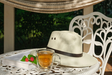 Transparent glass cup of herbal tea against the background of a white garden table and white sun hat in the garden. White book with raspberries and red currants. Shallow depth of field